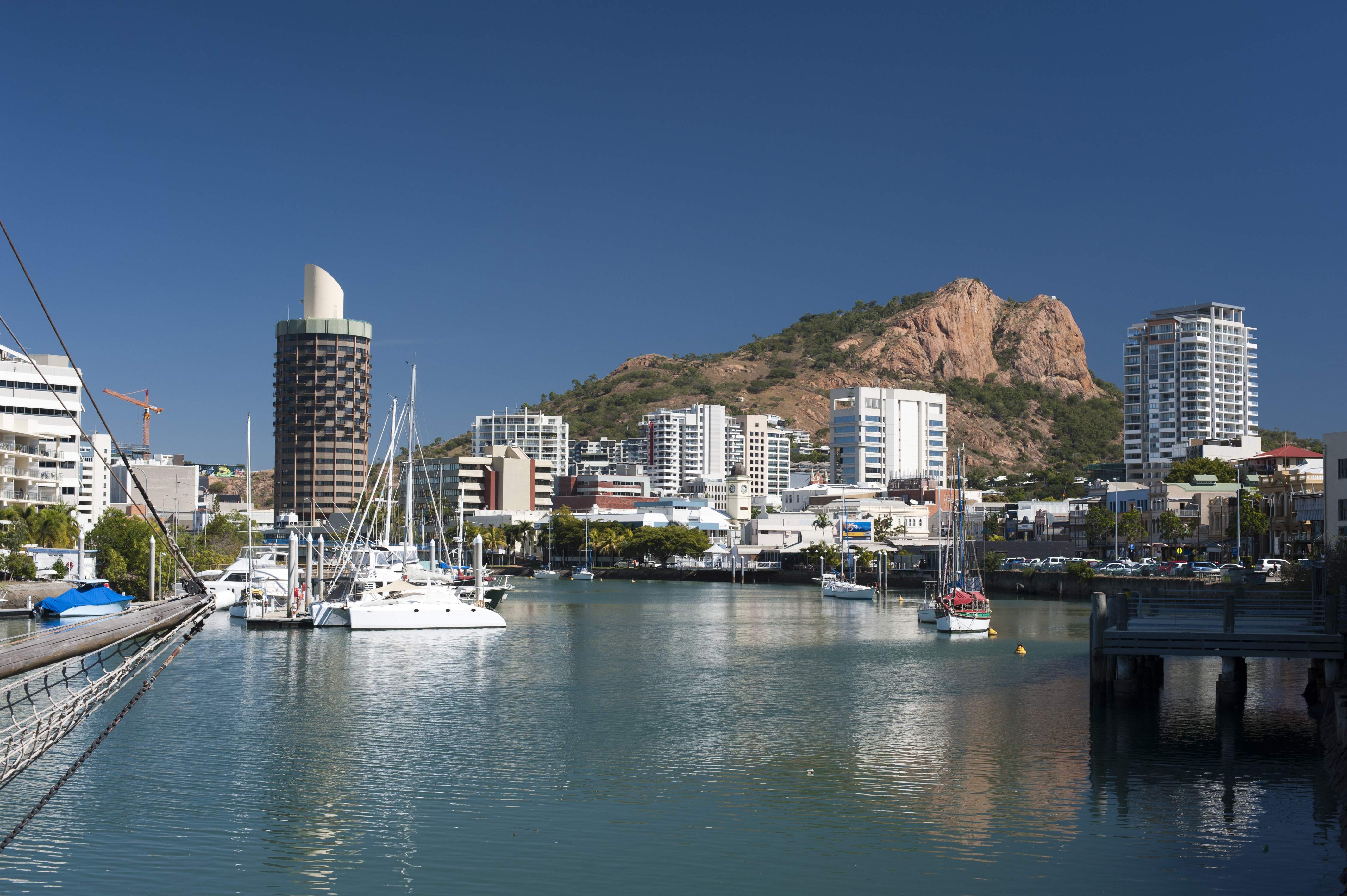 View Down The Quay Of Boats Moored In The Calm Water Of Townsville Marina In Queensland, Australia With Modern Waterfront City Central Architecture And A Castle Hill Behind